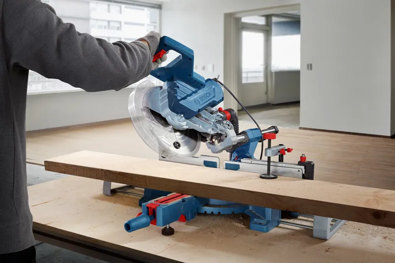 A person wearing safety equipment cuts a wooden plank using a miter saw.