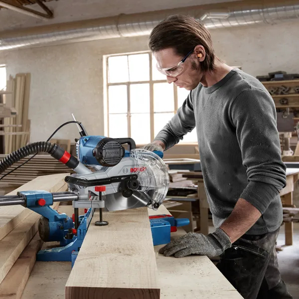 A person wearing safety equipment uses a miter saw to cut a wooden board in a workshop.