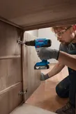 A person wearing safety equipment installs hinges on a wooden cabinet with a cordless drill.