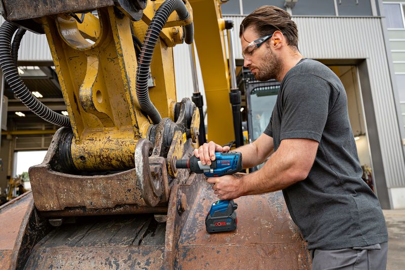 A person wearing safety equipment uses an impact wrench on heavy machinery outdoors.