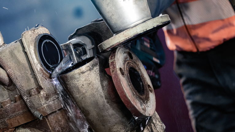 A person wearing safety equipment uses a grinder to cut a metal pipe with water cooling.