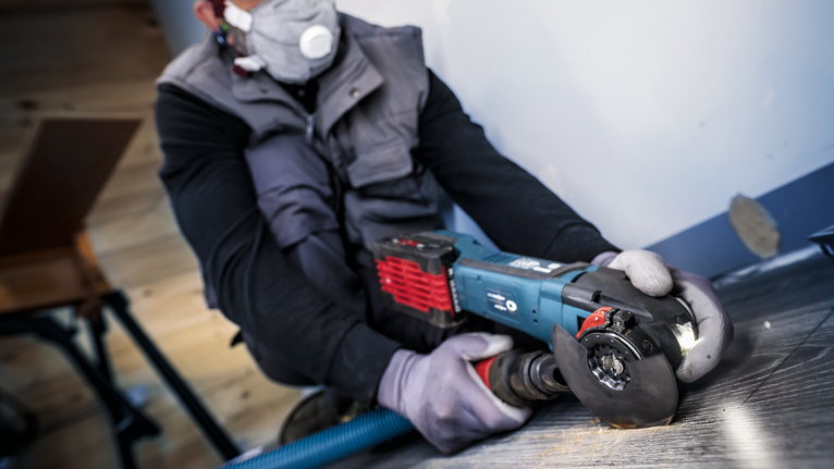 Person wearing safety equipment uses an angle grinder to cut flooring near a wall.