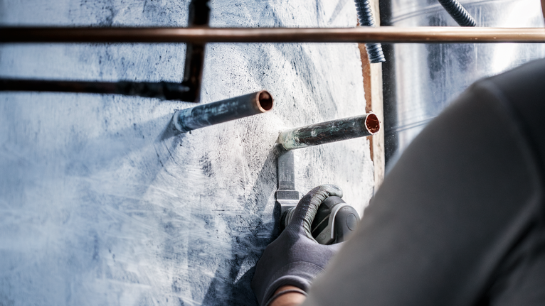 Person wearing safety equipment uses a power tool to cut into a wall around metal pipes.