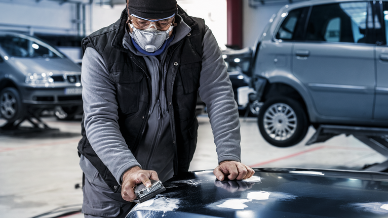 Person wearing safety equipment sands a car hood in an auto repair shop.