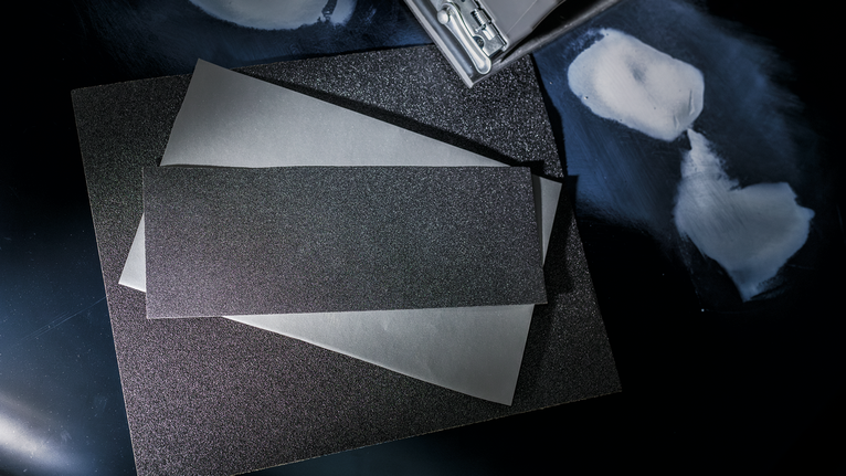 Sheets of sandpaper stacked on a dark surface next to areas prepared for sanding.