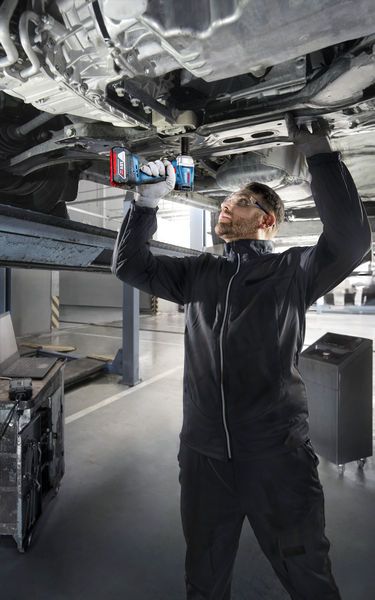 A person wearing safety equipment uses a power drill to work underneath a raised vehicle.