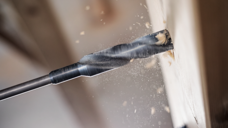 Large drill bit creating wood shavings while boring into a wooden board.