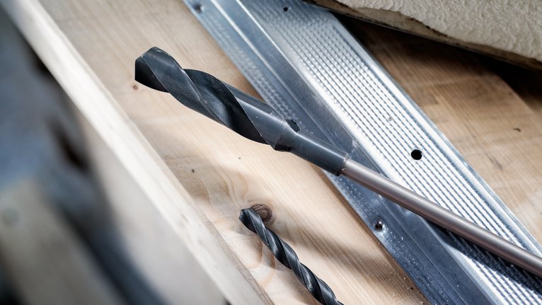 Large and small drill bits placed on a wooden surface near a metal guide.