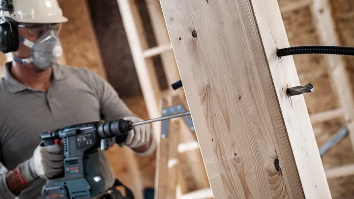 A person wearing safety equipment drills a large hole into a wooden beam.