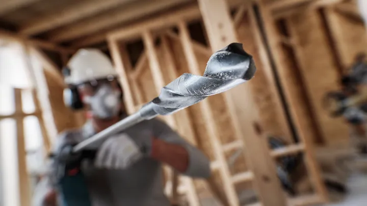 Person wearing safety equipment uses a large drill bit at a wood construction site.