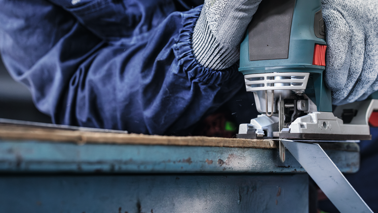 Person wearing safety equipment uses a jigsaw to cut a metal sheet.