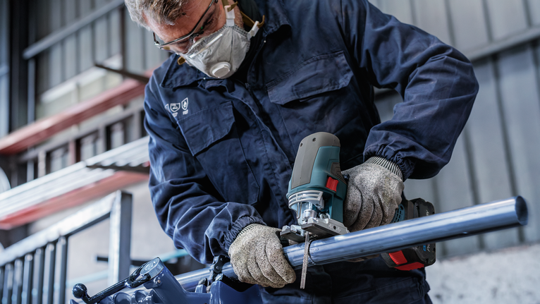 Person wearing safety equipment cuts a metal pipe with a jigsaw.