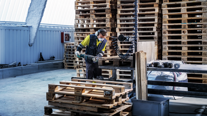 A person wearing safety equipment uses a power tool to cut wood pallets.