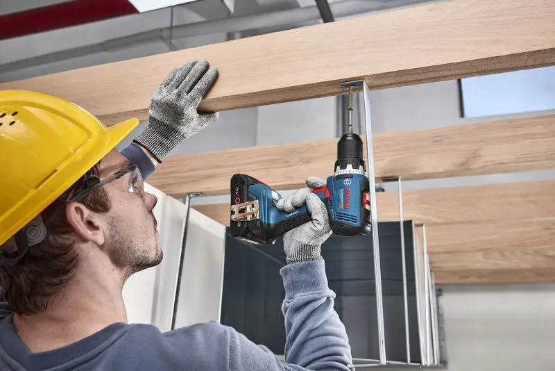 A person wearing safety equipment uses a cordless drill to fasten a screw into a wooden beam.