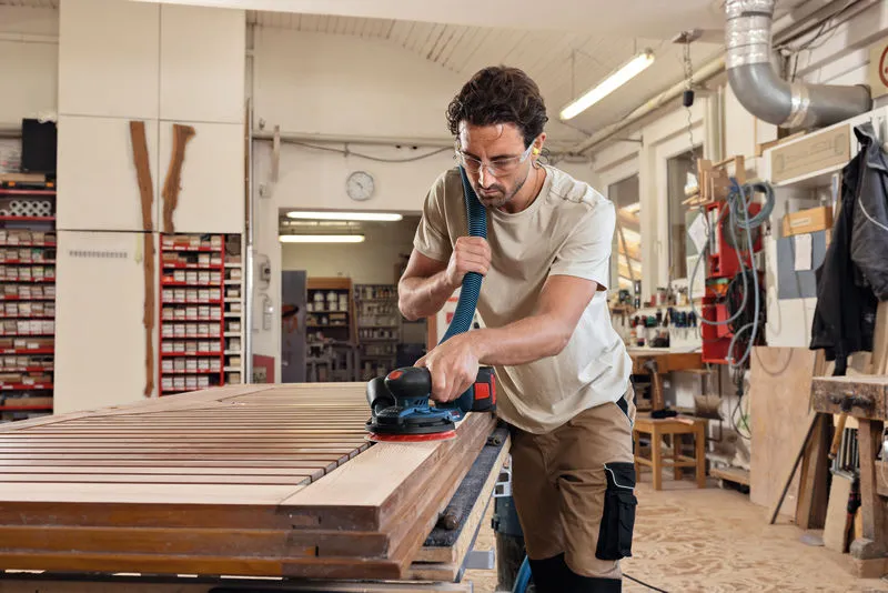 A person wearing safety equipment sands a wooden tabletop with a cordless random orbit sander.