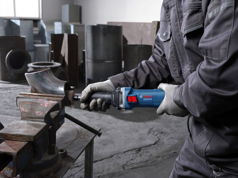 A person wearing safety equipment uses a straight grinder to grind a metal pipe in a workshop.