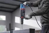 A person wearing safety equipment drills into a metal beam using a rotary hammer.