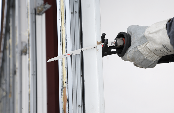 A worker wearing safety equipment uses a reciprocating saw to cut a window frame.