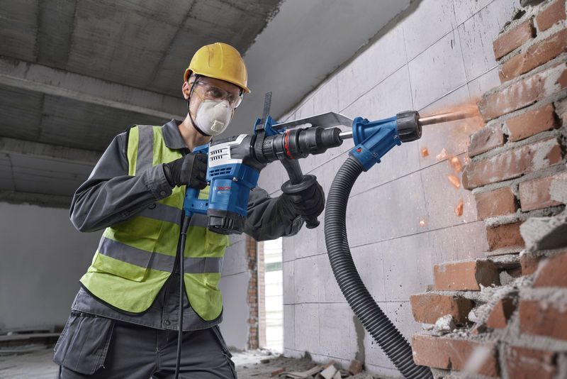 A person wearing safety equipment uses a demolition hammer with SDS max to break a brick wall.