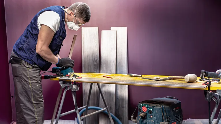 A person wearing safety equipment uses a power tool to cut wood on a workbench.