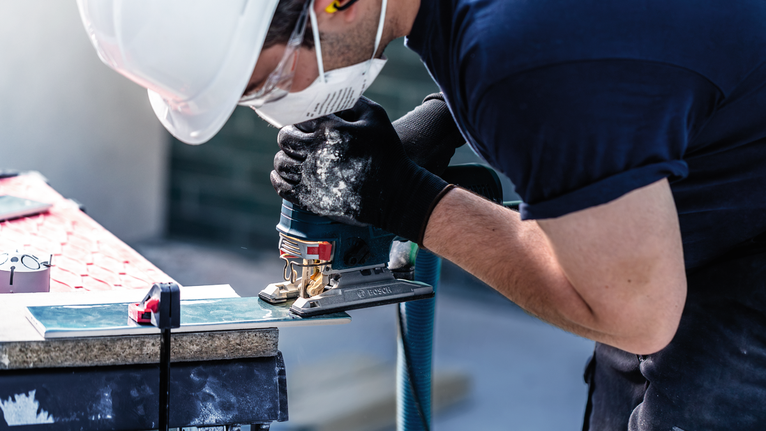 A person wearing safety equipment cuts a tile using a jigsaw.