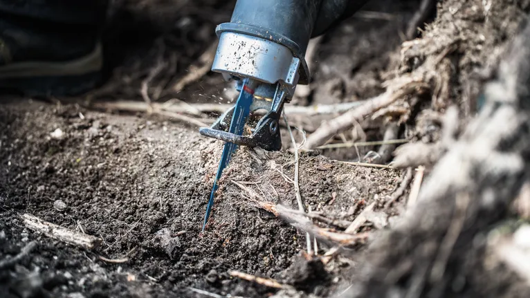 Person uses a reciprocating saw to cut roots in soil.