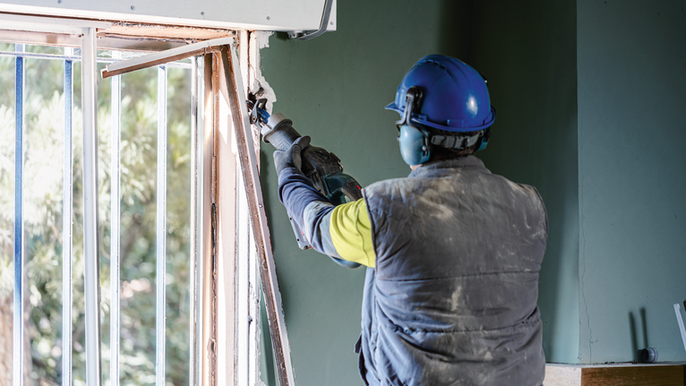 Person wearing safety equipment uses a power tool to cut or demolish a window frame.