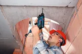 A person wearing safety equipment drills into a concrete ceiling using an impact drill.