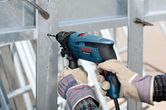 A person wearing safety equipment drills into a metal beam with an impact drill.