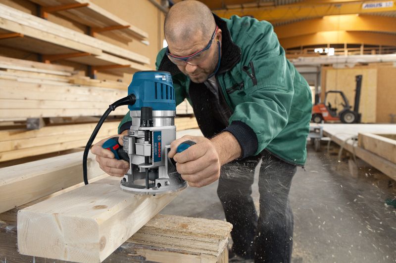 A person wearing safety equipment shapes a wooden beam using a multifunction router tool.