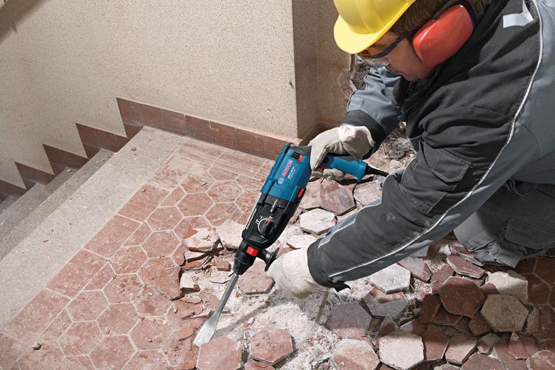 A person wearing safety equipment uses a demolition tool to break floor tiles.