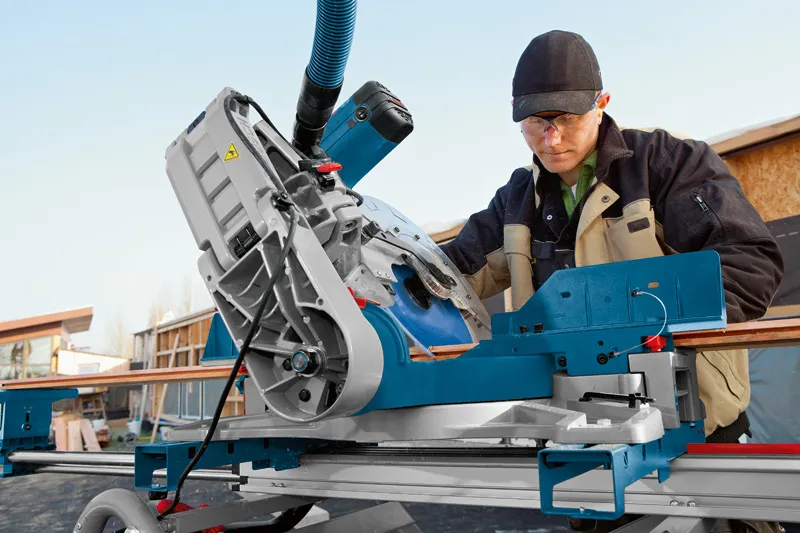 A person wearing safety equipment operates a mitre saw to cut wood on a jobsite.