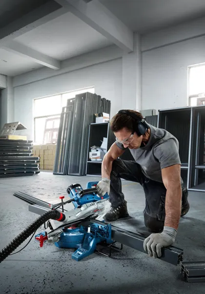 Una persona que usa equipo de seguridad opera una sierra ingletadora para cortar una viga de metal en el interior.