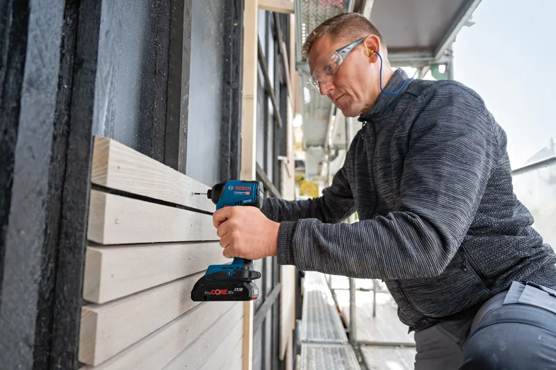 Una persona que usa equipo de seguridad utiliza un taladro inalámbrico para fijar tablas de madera al aire libre.