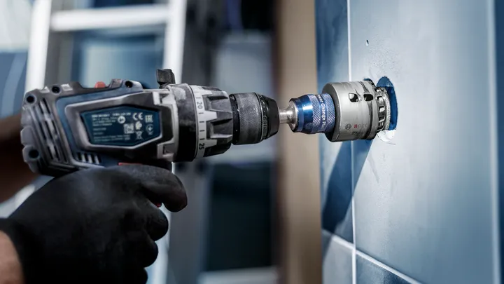 Una persona que usa equipo de seguridad utiliza un taladro eléctrico con una sierra de corona en una pared de azulejos.