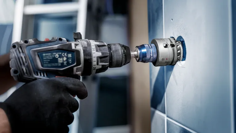 Una persona que usa equipo de seguridad utiliza un taladro eléctrico con una sierra de corona en una pared de azulejos.