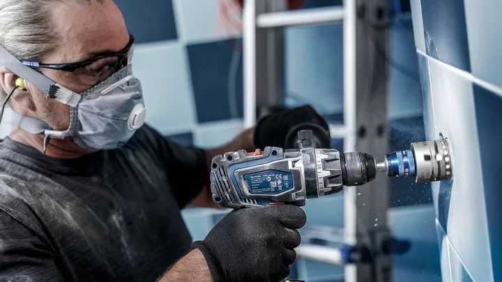 Una persona que usa equipo de seguridad perfora un agujero circular en una pared de azulejos.