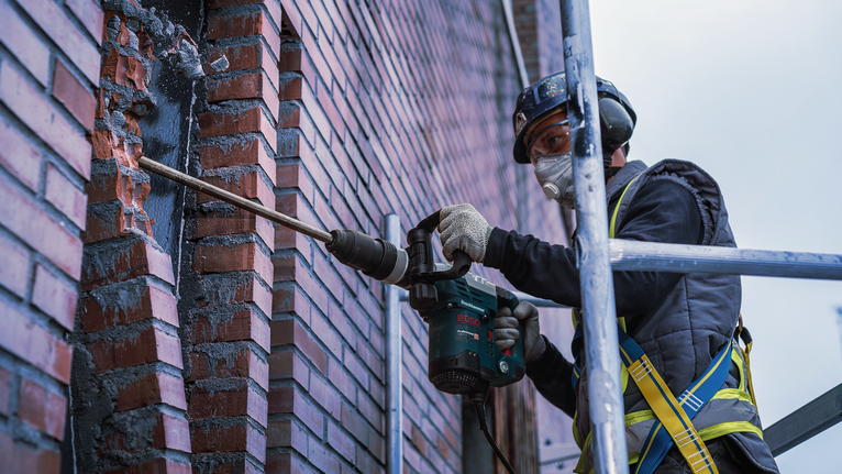 Una persona que usa equipo de seguridad utiliza un martillo rotatorio para romper una pared de ladrillos.