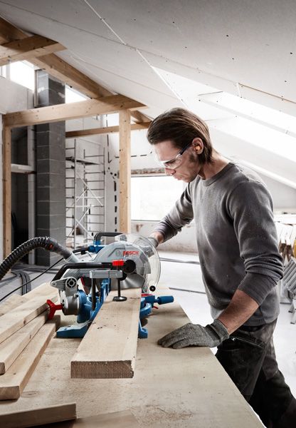 Una persona que usa equipo de seguridad corta tablones de madera con una sierra ingletadora deslizante.