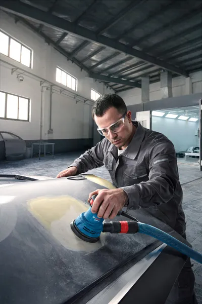 Una persona que usa equipo de seguridad lija el capó de un automóvil en un taller de reparación de automóviles.