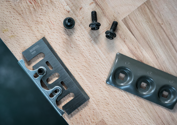 Hoja de metal, tornillos de fijación y placa de montaje dispuestos sobre una superficie de madera.