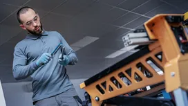 Una persona que lleva equipo de seguridad inspecciona dos herramientas metálicas junto a una estructura industrial de color naranja.