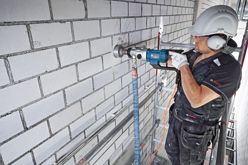 A person wearing safety equipment drills into a brick wall with a diamond drill.