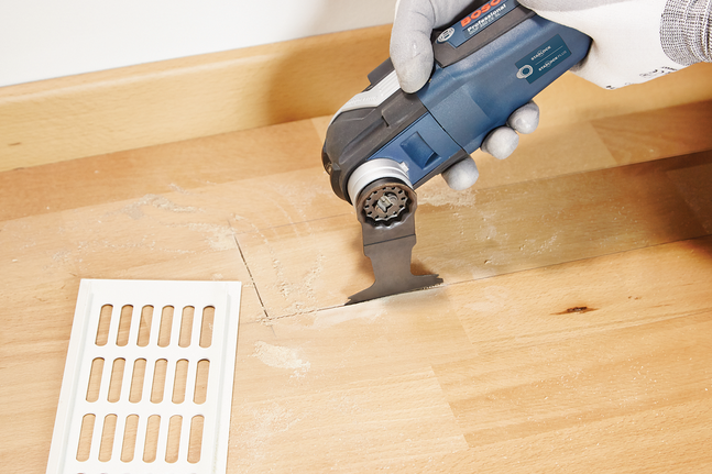 Person wearing safety equipment cuts a floorboard with a multi-tool near a floor vent.