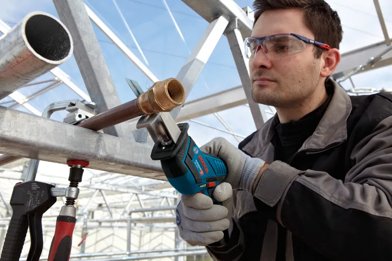 A person wearing safety equipment cuts a metal pipe with a reciprocating saw.