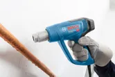 A person wearing safety equipment uses a blue heat gun near a copper pipe.