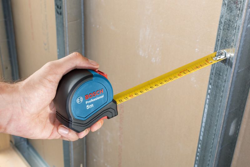 A person uses a tape measure to check distance between drywall and metal frame.