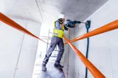 A person wearing safety equipment drills into a concrete ceiling with a rotary hammer.