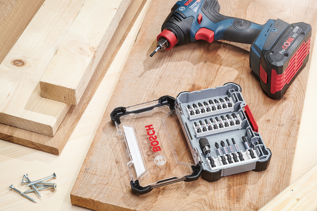Cordless drill and bit set on a wooden surface with screws nearby.