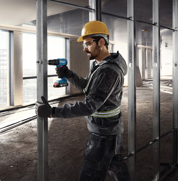 A person wearing safety equipment drills into a metal stud in a building under construction.